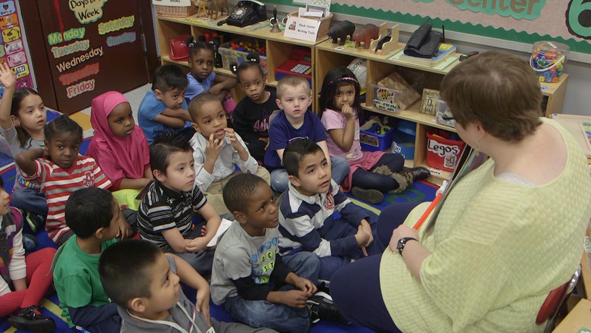 Picture of children sitting on the floor listening to teacher read book.