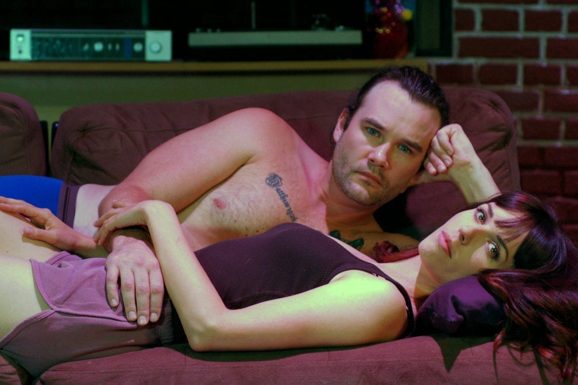 A man and woman laying on a sofa as part of a play, "The Way We Get By."