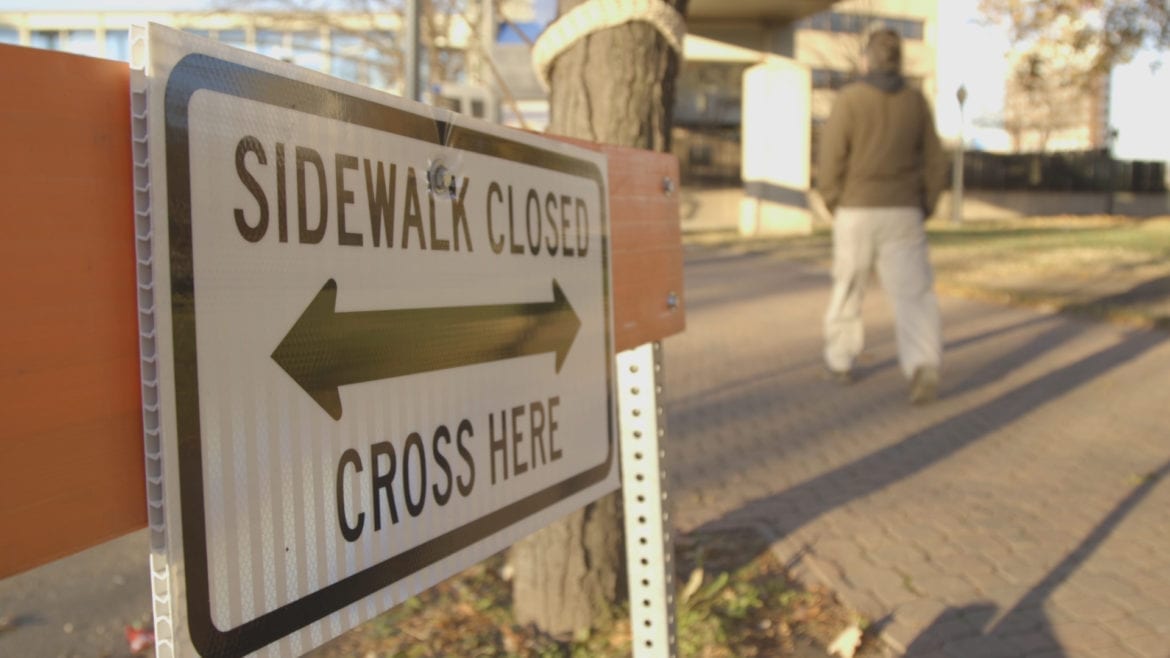 A man walking on a sidewalk near a "Sidewalk Closed, Cross Here" sign.