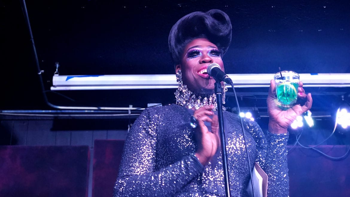 Moltyn Decadence, a drag queen in Kansas City, holds a green drink behind the mic while smiling, in her bouffant and sequin gown