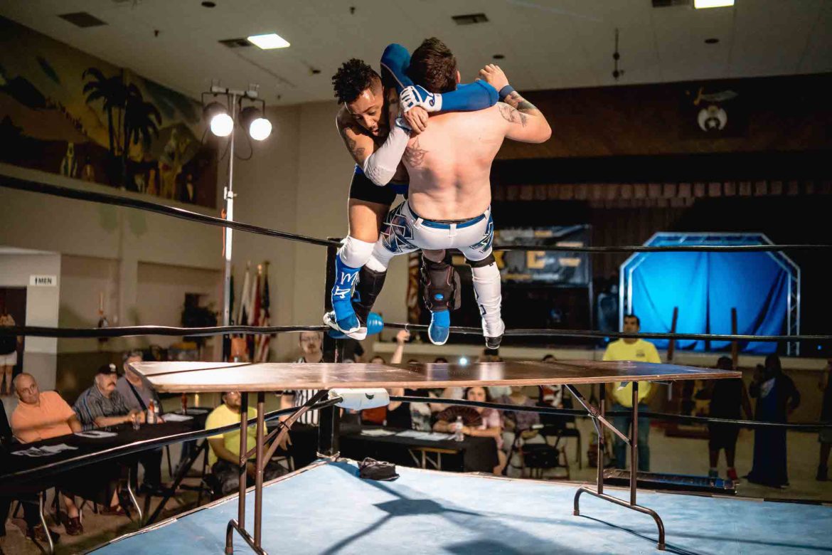 A wrestler takes another wrestler off the turnbuckle and through a table.