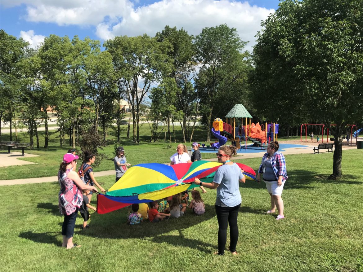 staff and children at summer camp activity under a parachute at a park