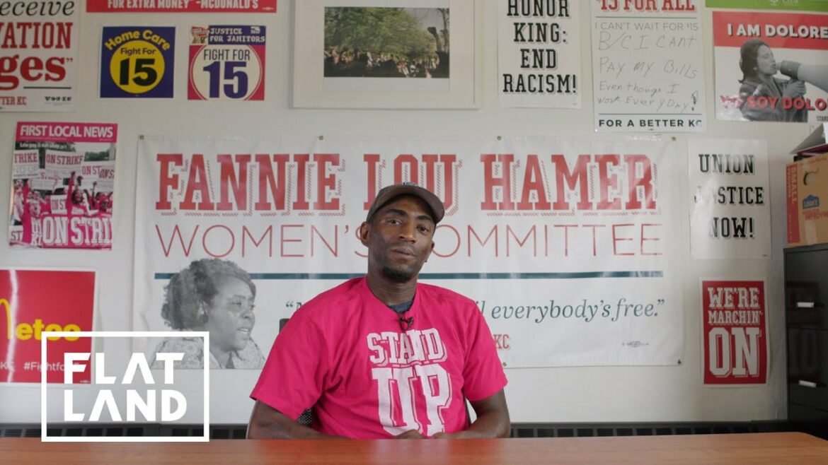 Man in red shirt that reads "stand up" sits at table with Fannie Lou Hamer board behind him.