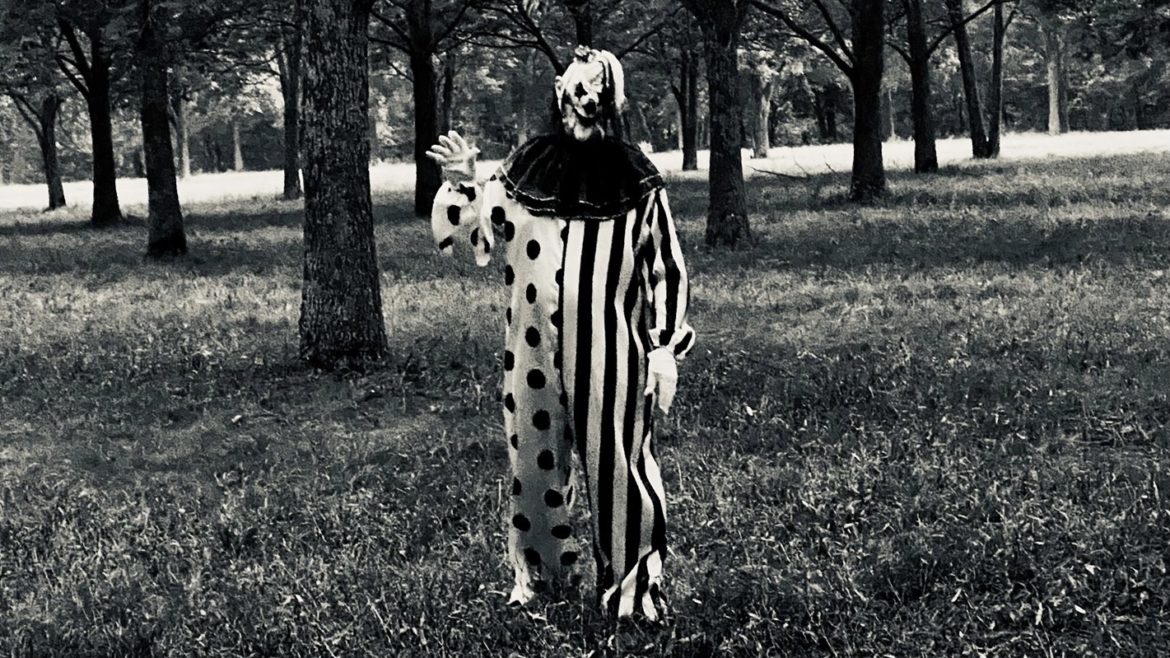 A clown stands in a field in a creepy outfit.