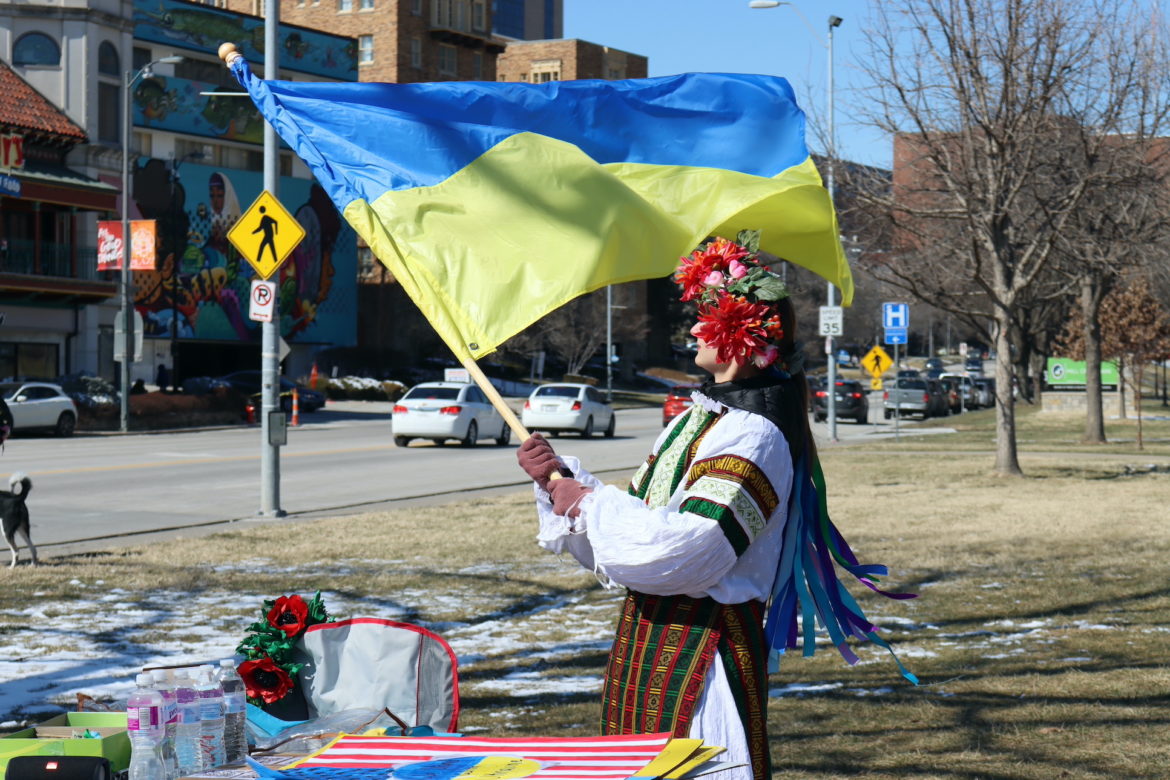 Woman in flower crown and traditional, colorful Ukrainian dress holds a blowing Ukrainian flag in a park.