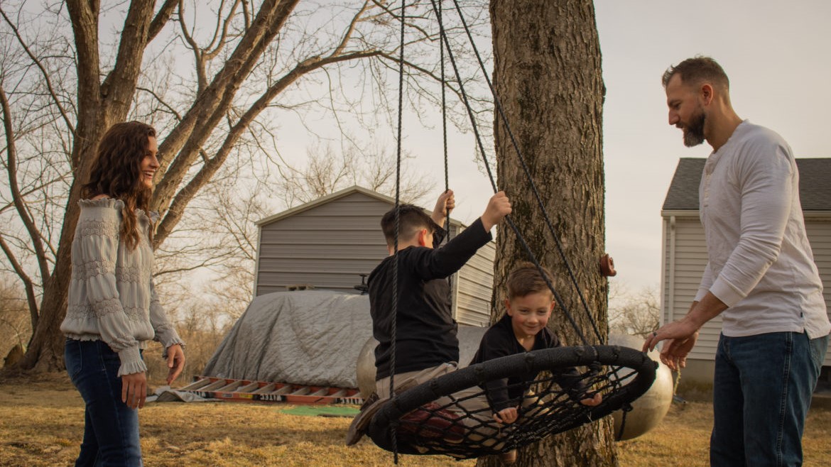 Woman and man push two young boys on a rope swing.