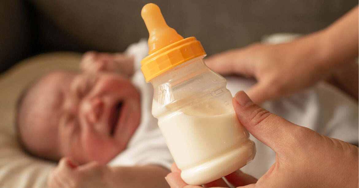 A baby bottle is held up as a baby cries in the background, illustrating the difficulty parents are having during the formula shortage. Image by Adobe Spark.