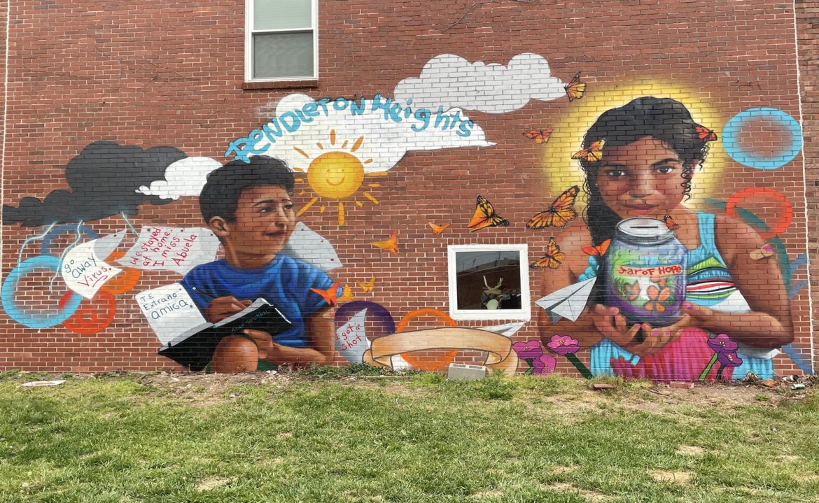 A mural of two young, Brown children painted on a red brick wall illustrates the difficulty kids felt being isolated from friends and family during the pandemic. One of the images shows a boy holding a notebook, with pages flitting around him that say "Te extraño amiga" and "go away virus."