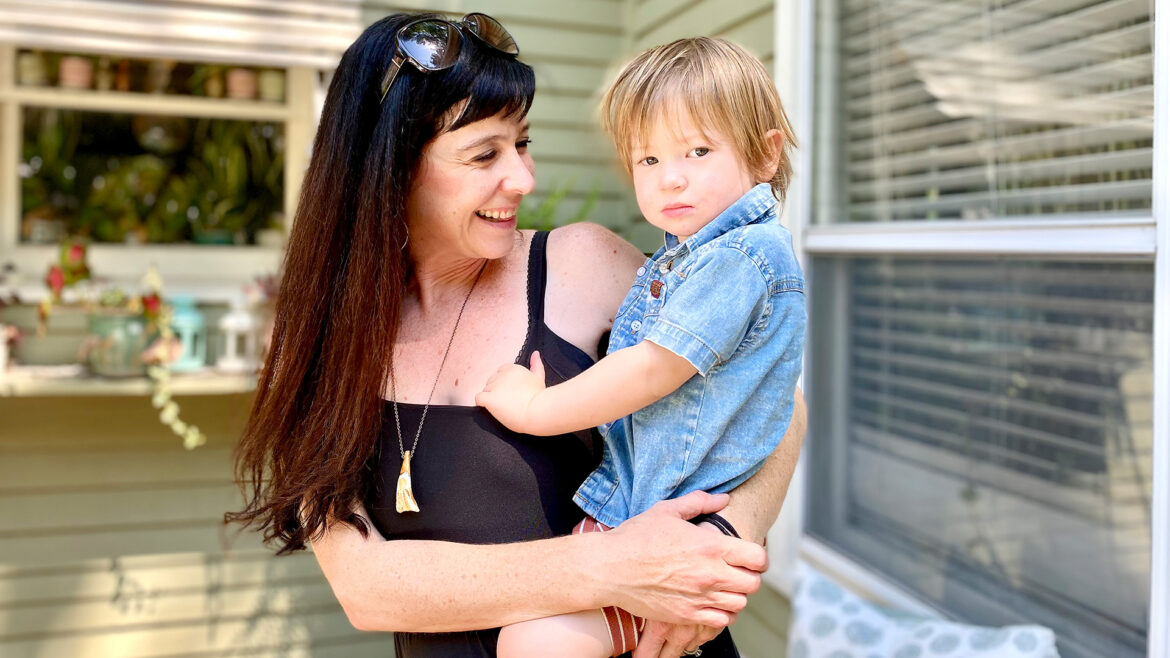 A mother with long brown hair holds her son, who is around two years old, outside of their green home. Lisa Sauciuc was recently able to get her youngest vaccinated from COVID-19.