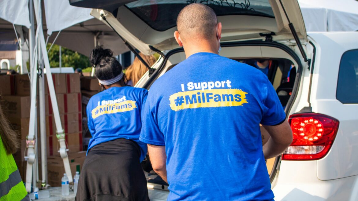 Two people wear shirts that say "I support Mil Fams" and load food into the back of a vehicle.