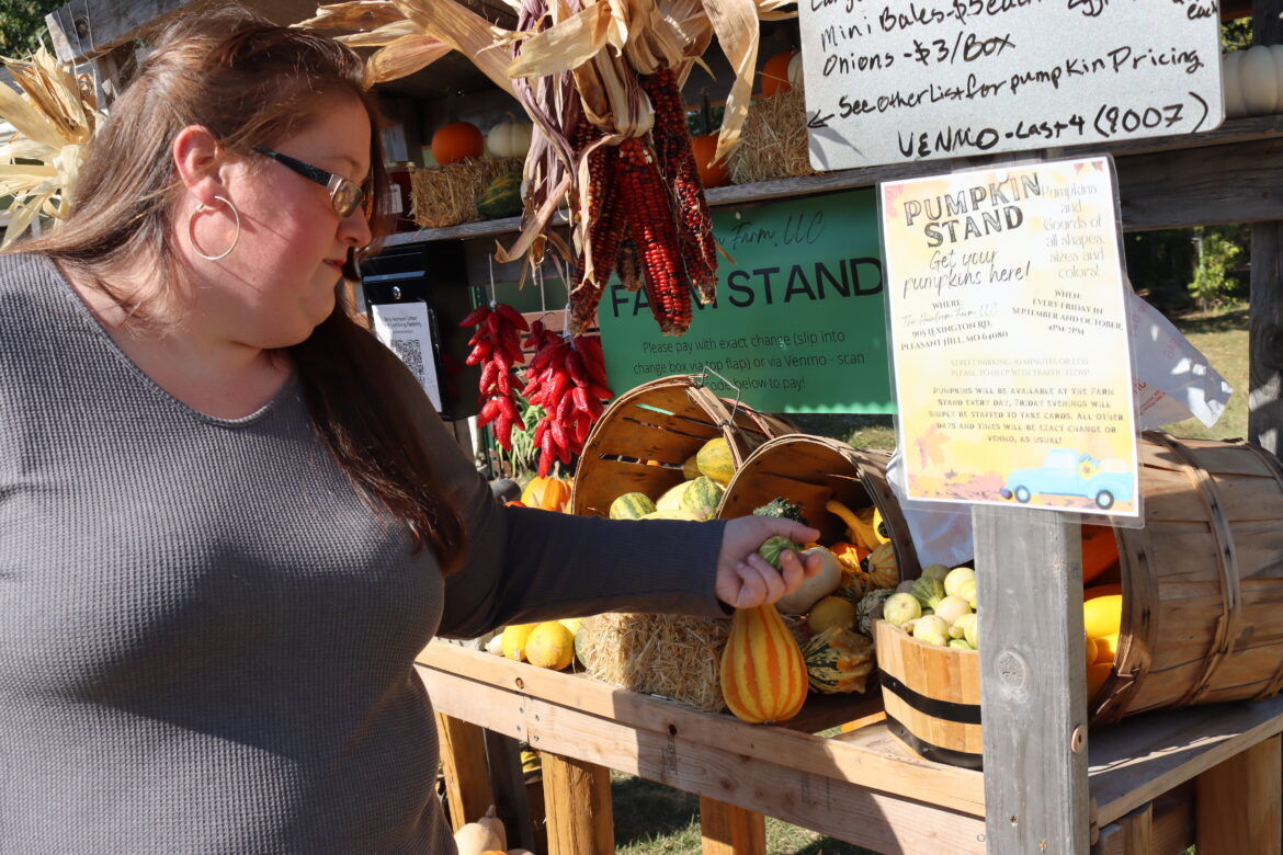 woman in gray shirt and glasses points to squash in a wooden farm stand.