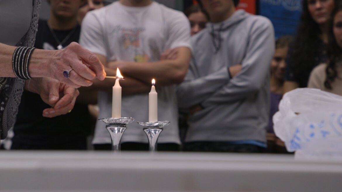 A elderly woman holds a match over a white candlestick. She's wearing a large amethyst ring. High schoolers watch in the background.