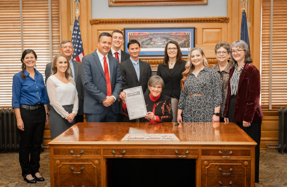 Group of people stand around KS Governor Laura Kelly at her desk. Kelly holds a document proclaiming rural national health day in Kansas.
