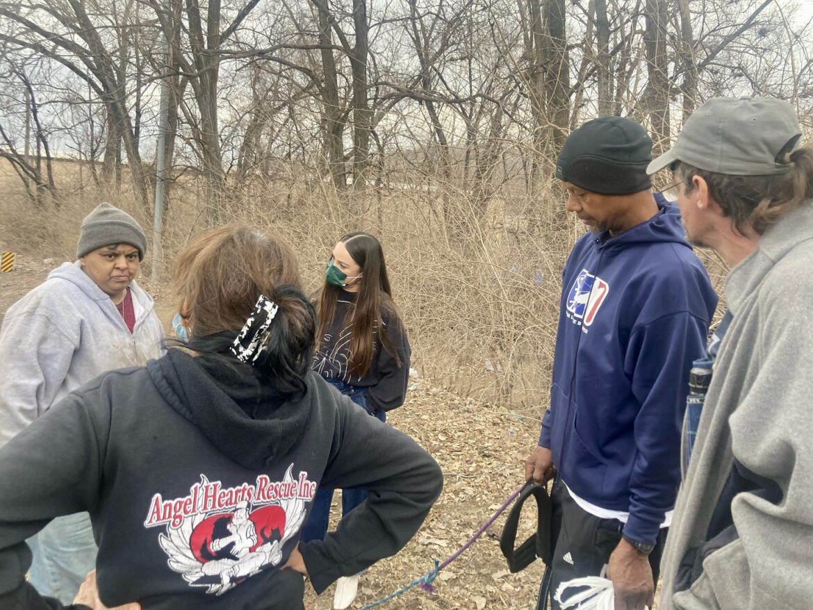 A group of people stand outside. Several people belong to an encampment where people experiencing homelessness live. One woman Alice Delbosque has her back to the camera. She is wearing a sweatshirt of her organization called Angel Hearts Rescue, which provides aid to pets belonging to those experiencing homelessness.