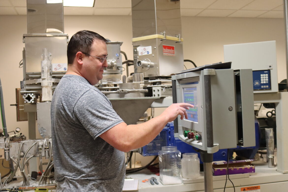 Man in a grey shirt and glasses uses the touchscreen on a large machine used for extruding plastic pellets.