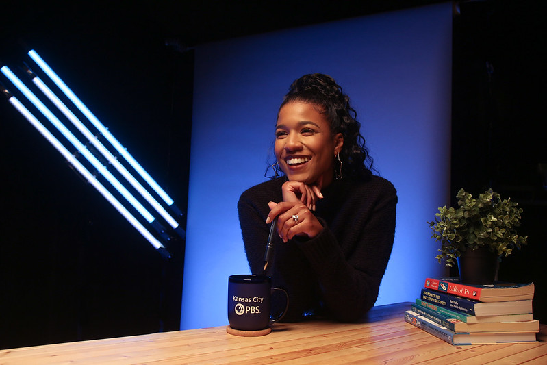 woman sits in front of a blue background. She has a coffee cup that reads "Kansas City PBS" and a stack of books with a plant next to here.
