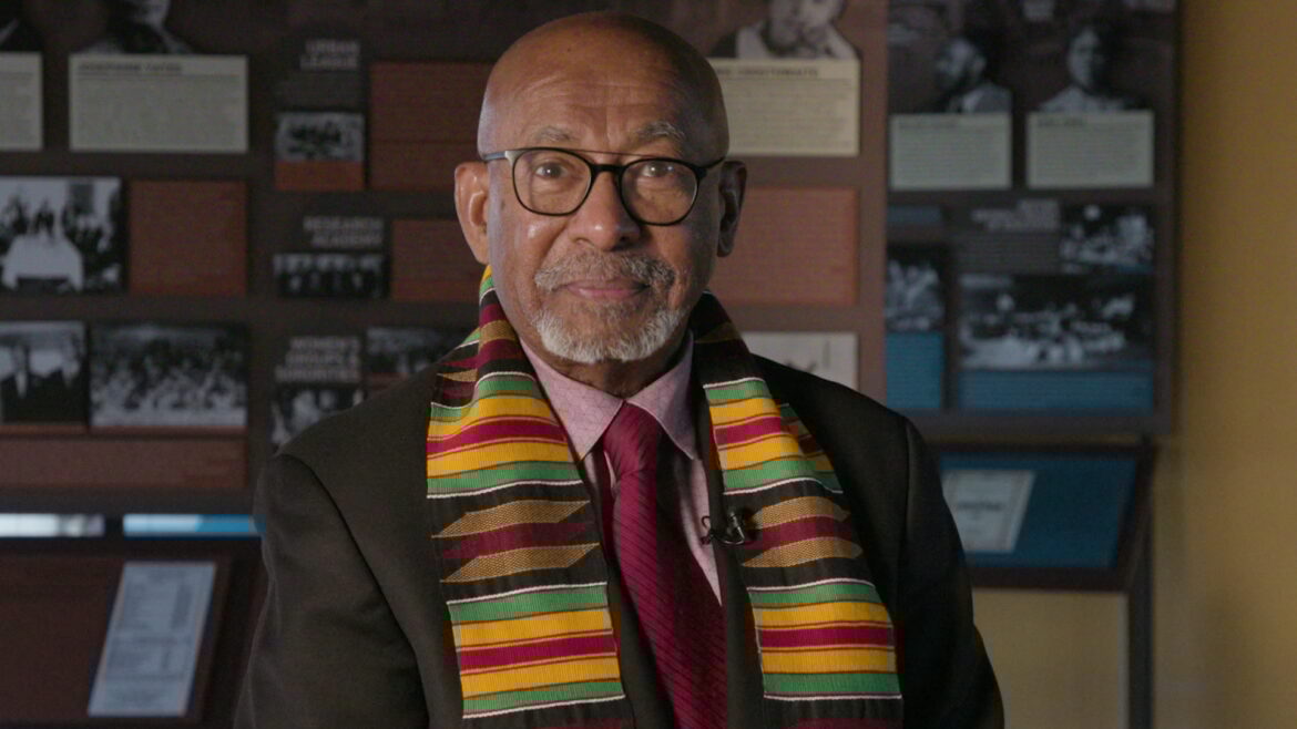 A man with white facial hair and classes sits in front of a display with historical documents. He wears a traditional scarf with reds, yellows, blacks and greens.