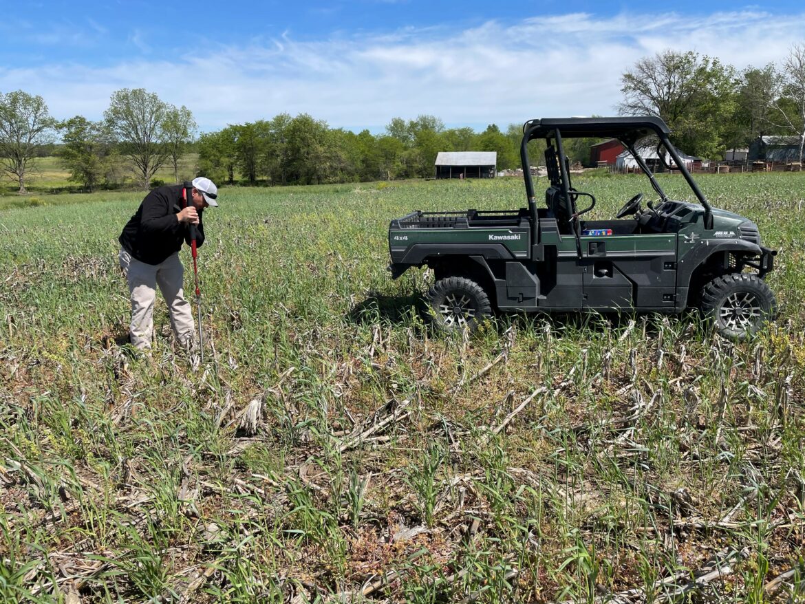 A man stands in a field that has dead corn stalks and a young cover crop. He is digging for a soil sample. Next to him is a green four wheeler.