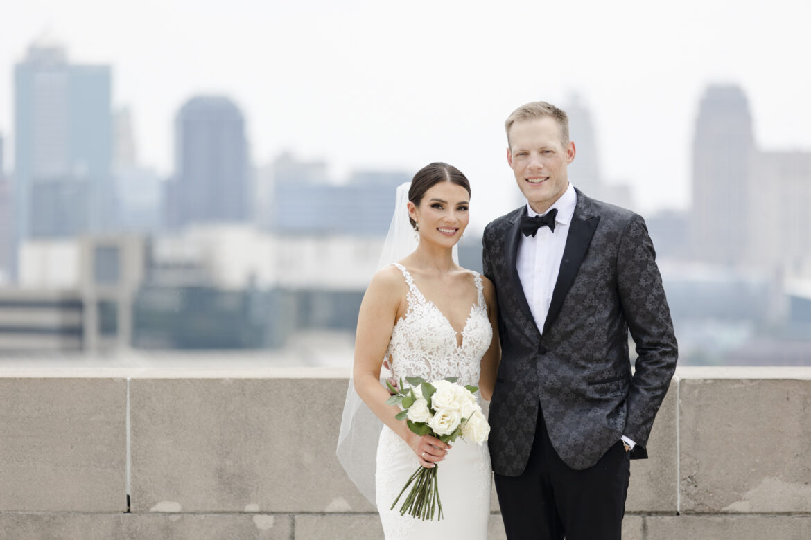 A wedding portrait. A young white couple, woman wearing a white gown holding a bouquet, and man in a tuxedo, stand outside of Kansas City's citscape. Joey and Audrey Sughroue got married July 2022. Their hope this year is to find a living donor for Joey, whose kidneys are at 10% function. (Contributed image)
