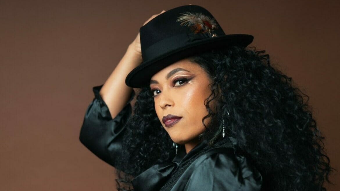 A woman with long curly hair, in a black silk shirt and black fedora hat looks into the camera.