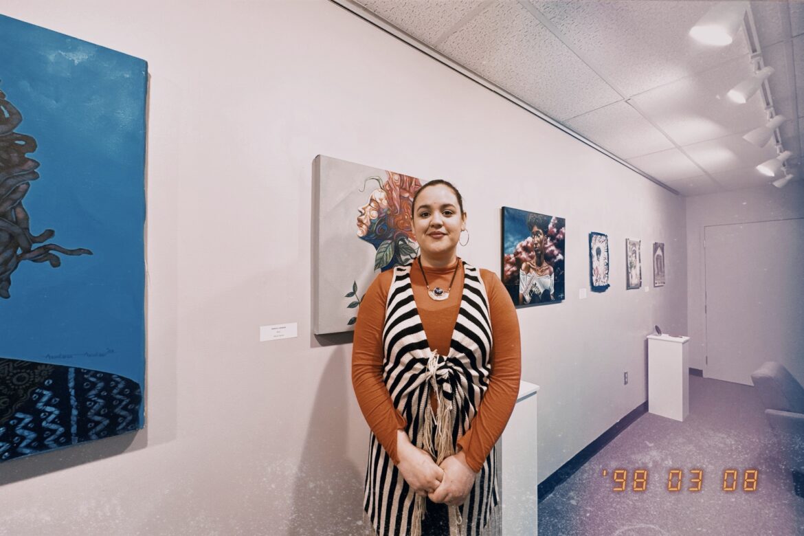 A woman wearing a striped shirt over an orange long sleeve shirt poses in an art gallery setting. Shai Perry is gallery coordinator at the Kansas City Kansas Community College. She is also an artist and is from Tennessee. Photo by Vicky Diaz-Camacho for Flatland.