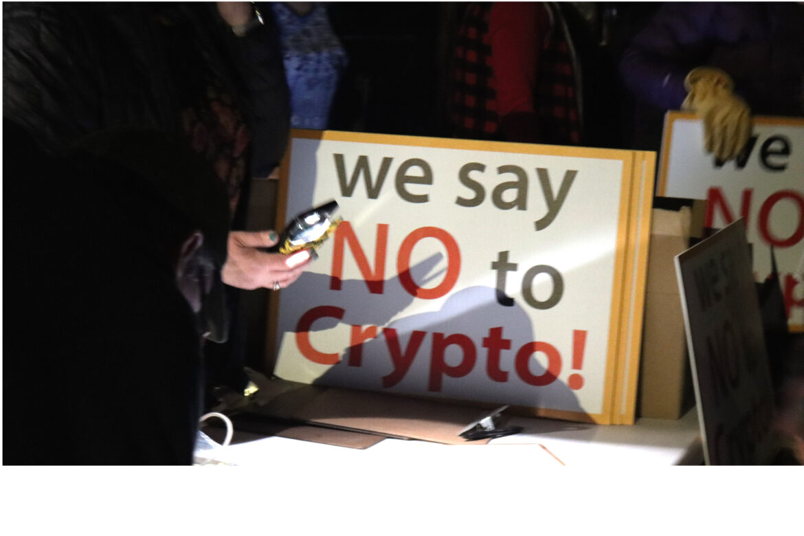 someone holds a phone with the flashlight on up to a table. Next to them sits a stack of signs that read "we say NO to Crypto"