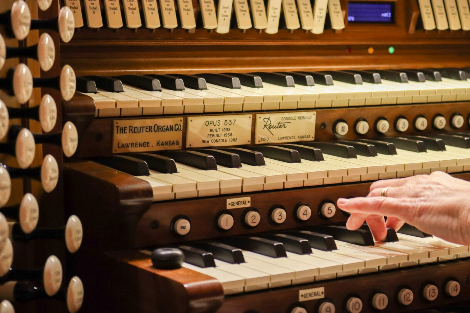 Between rows of piano keys (stacked vertically) we see plaques that read "Reuter Organ Company" A hand reaches to play several keys.