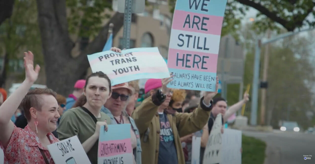 Folks hold up pink white and blue signs that read "We Are Still Here" at a transgender rights protest in Kansas City.
