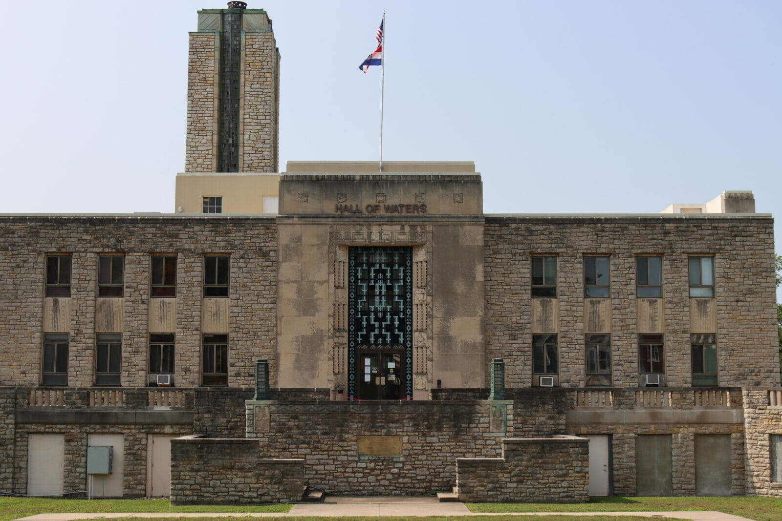 A stone building with bluish green metal on the front reads "Hall of Waters" a stone water tower is on top of the building, as is a flag.