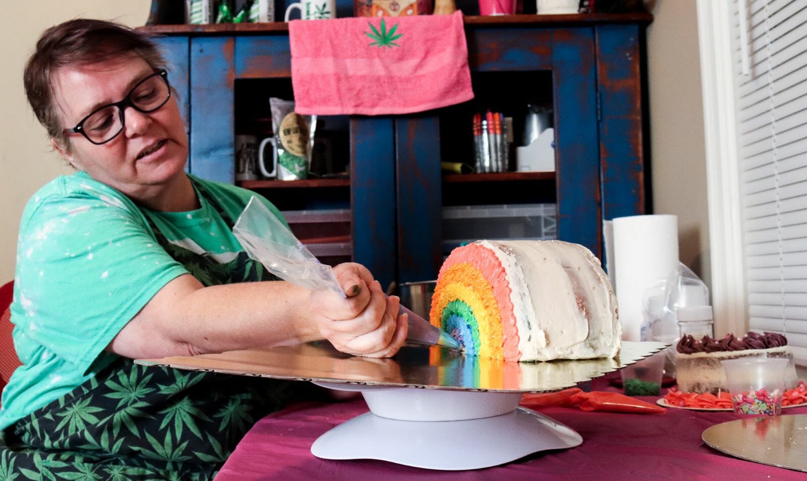 a woman with glasses and an apron covered in marijuana leaves holds a piping bag to a cake decorated to look like a rainbow.