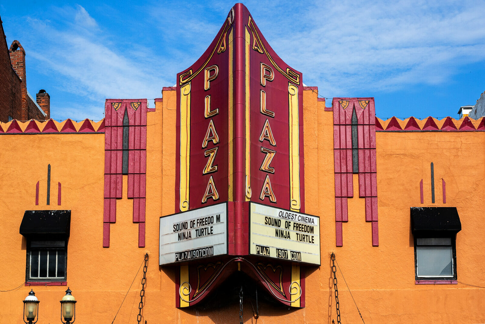 An orange and red building facade shows a big red movie theatre sign that reads "Plaza" on the marquee are movies playing at the theatre and a note reading "oldest cinema"