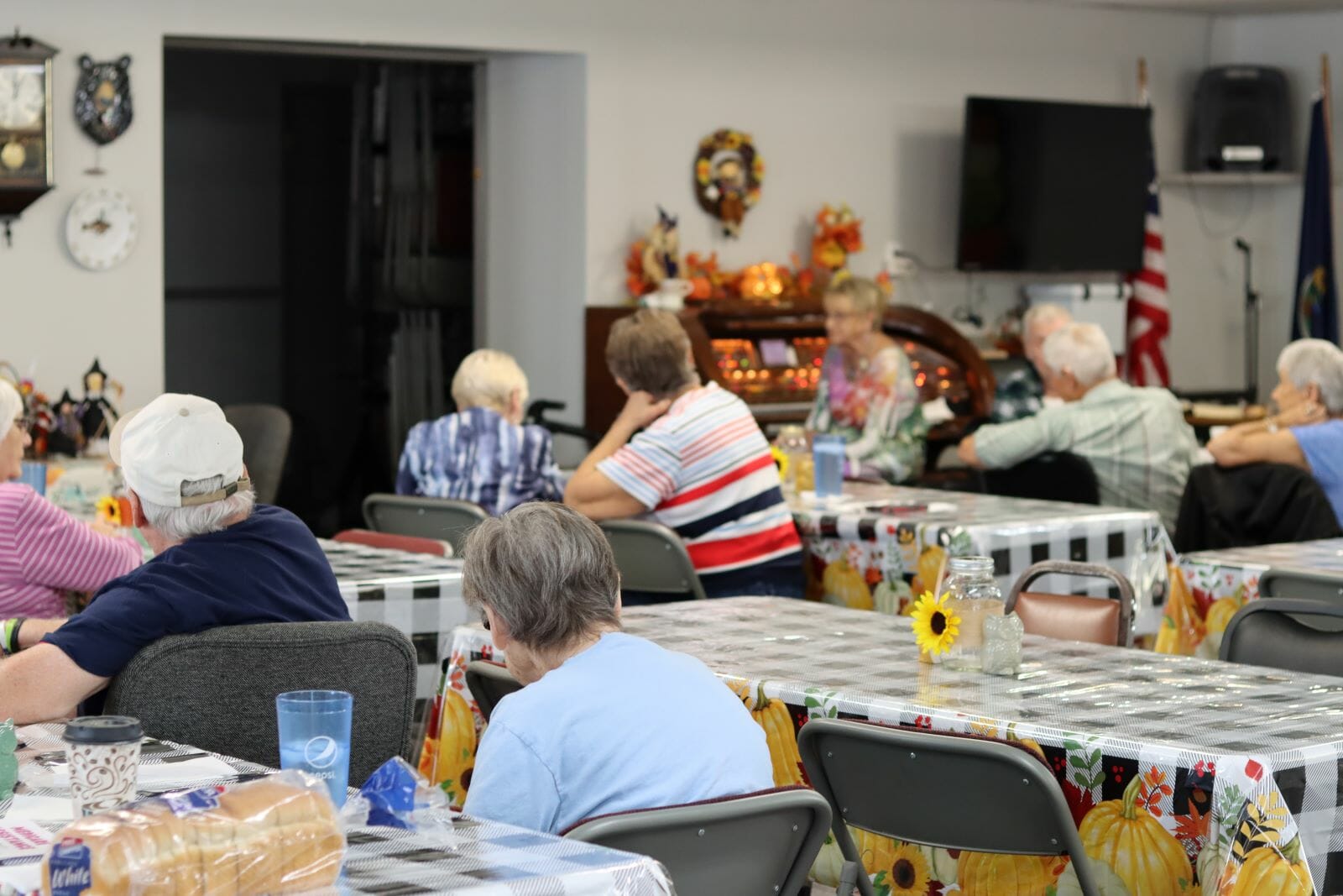 A group of older people sit at rectangular tables with checkered table cloths. In the background a woman sits at an electric organ with lots of colorful lights.