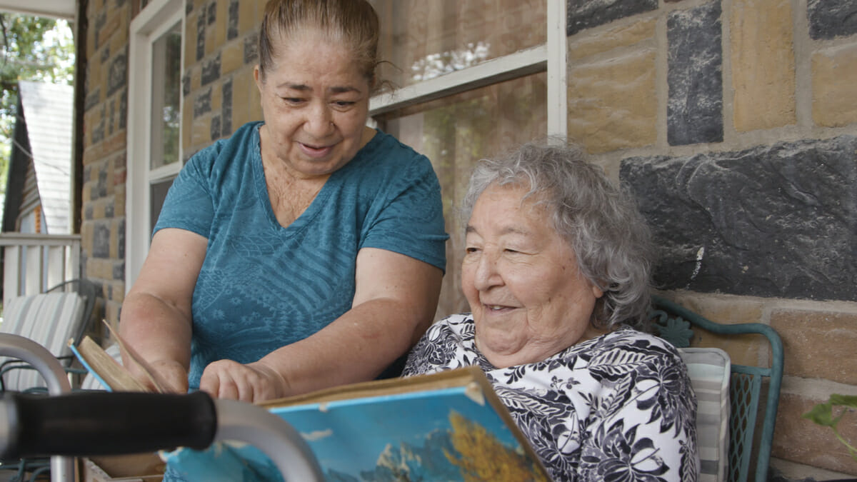 Woman sitting with older woman on porch, reading a book.