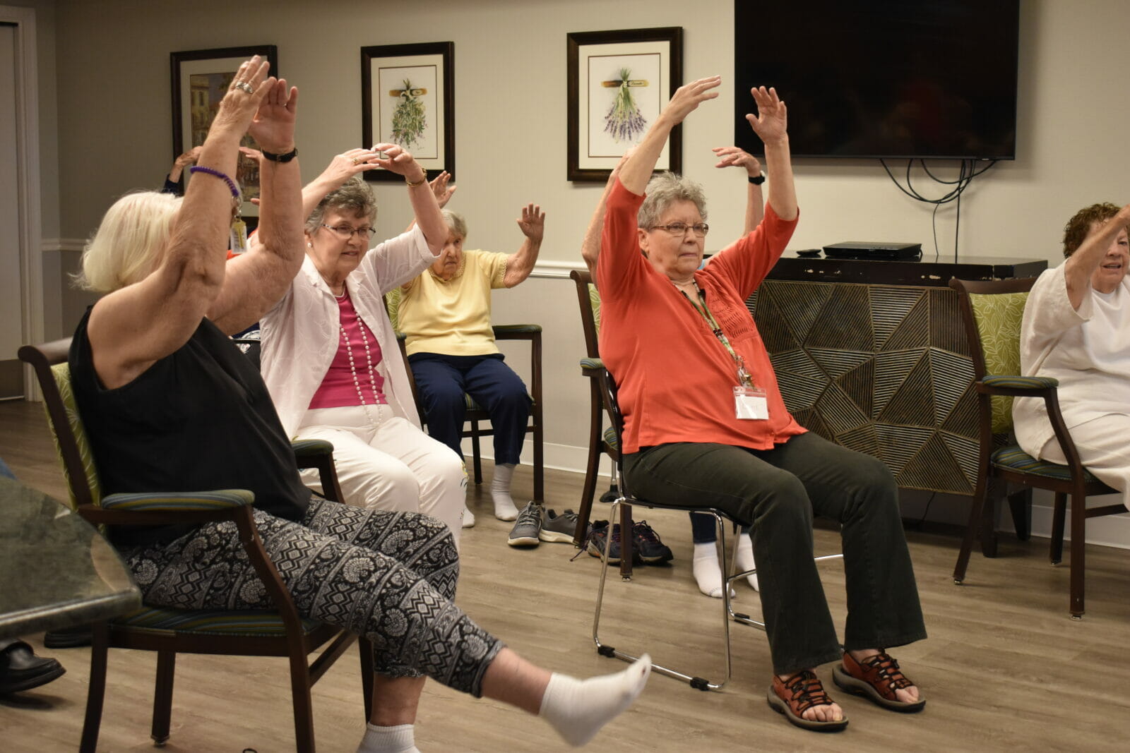 A group of older women hold their arms up in fifth position while seated.