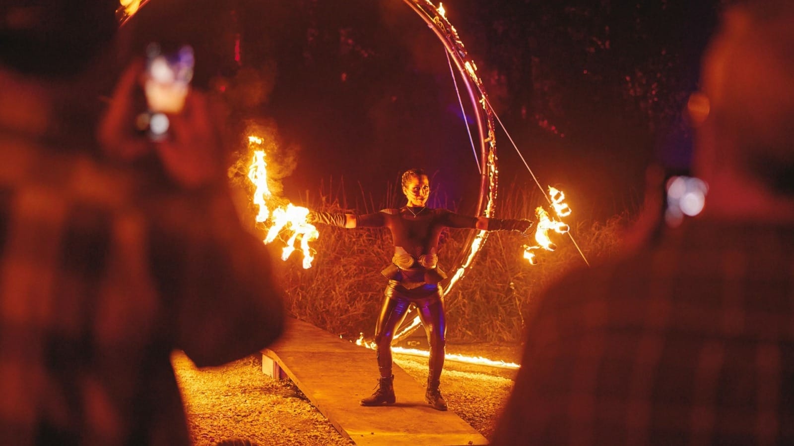 A fire performer in black stands waving flames in the dark forest.