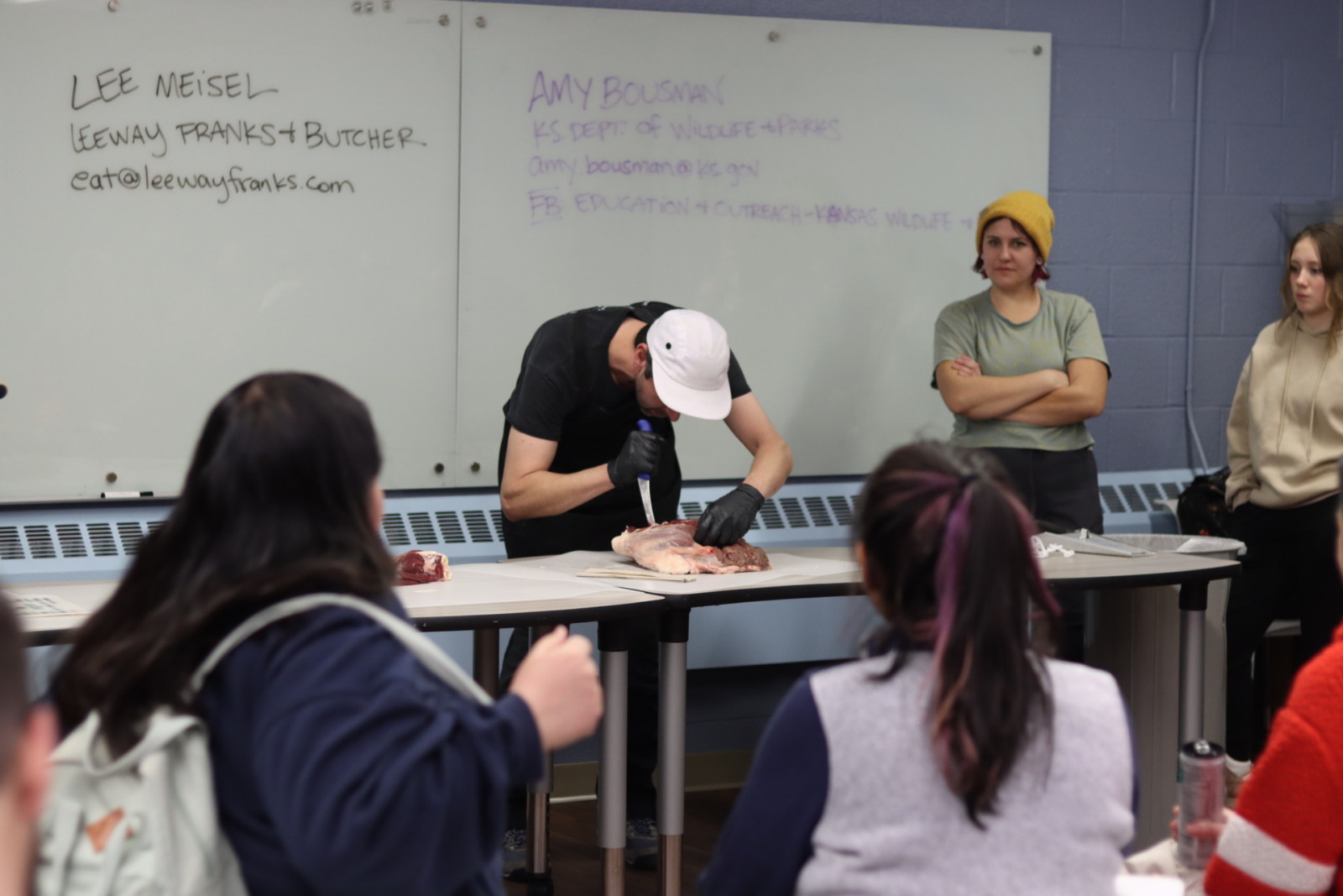 Man holding knife bends over a table cuts into slab of raw meat. People in a classroom crowd around to watch.