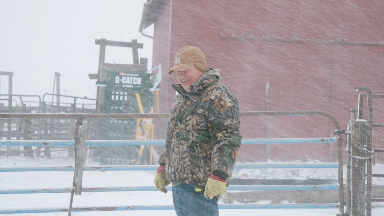 A man in a camouflage coat and a brown hat stands in front of a red barn in a blizzard.