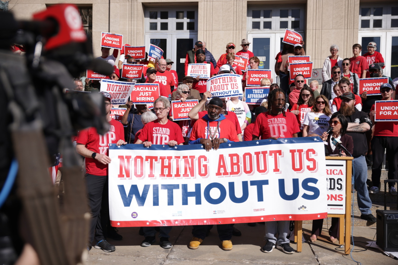 Group of activists in red T-shirts hold banner reading Nothing About Us Without Us