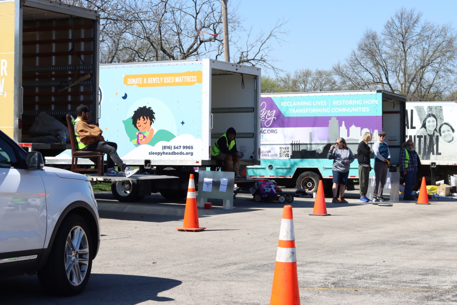 A row of box trucks with various nonprofit logos are lined up in a parking lot. A car drives down a row of cones behind the trucks while volunteers stand nearby in yellow vests.