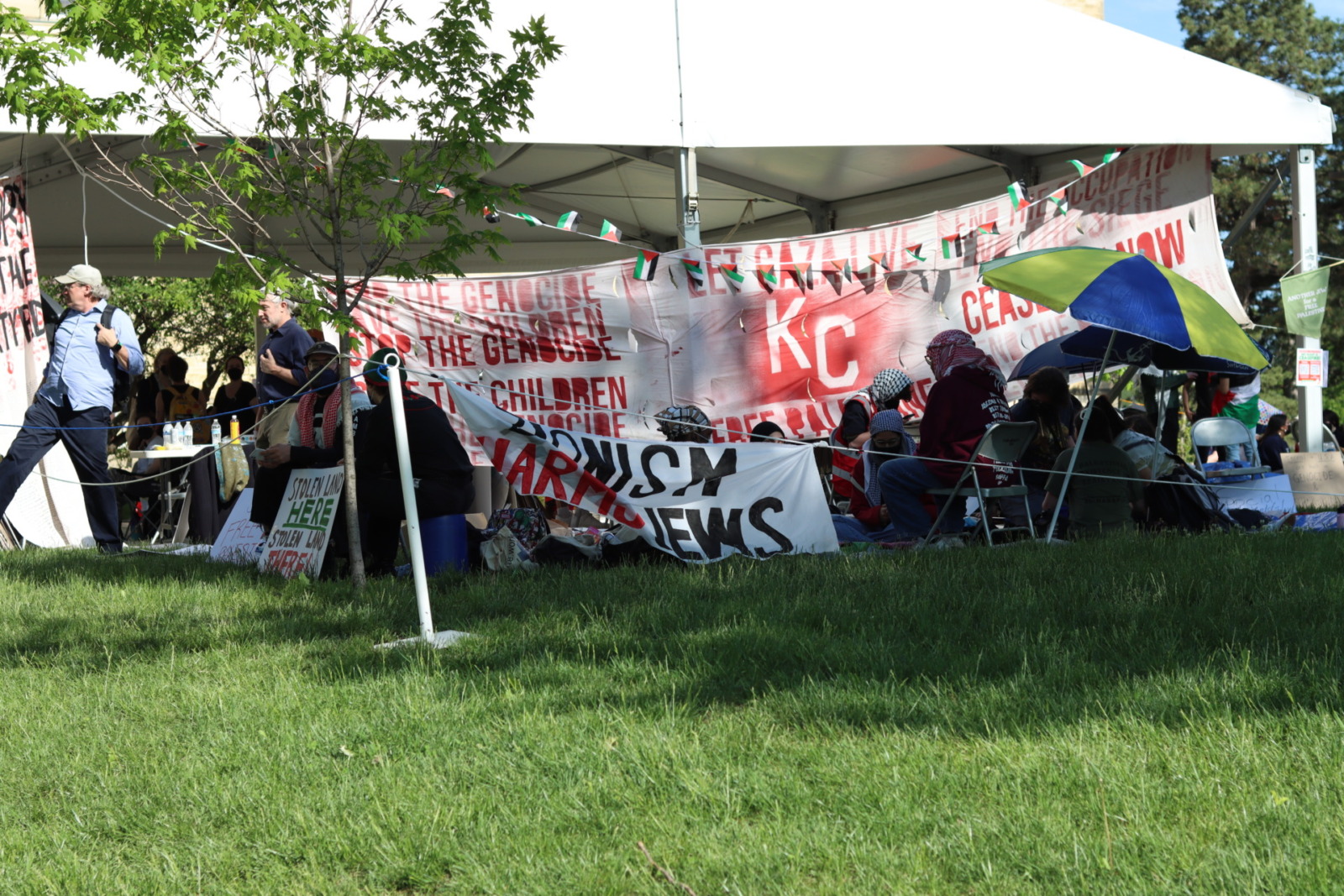 A large white tent is covered with banners reading things like "stop the genocide" "Ceasefire now" "Let Gaza Live". Around the tent sit students wearing keffiyehs.