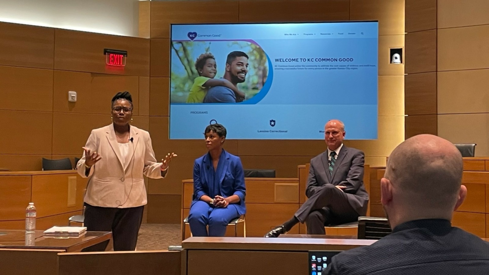 A man, back turned, faces a panel of three people. One woman is standing, and a man and a woman are seated. A large screen behind them.