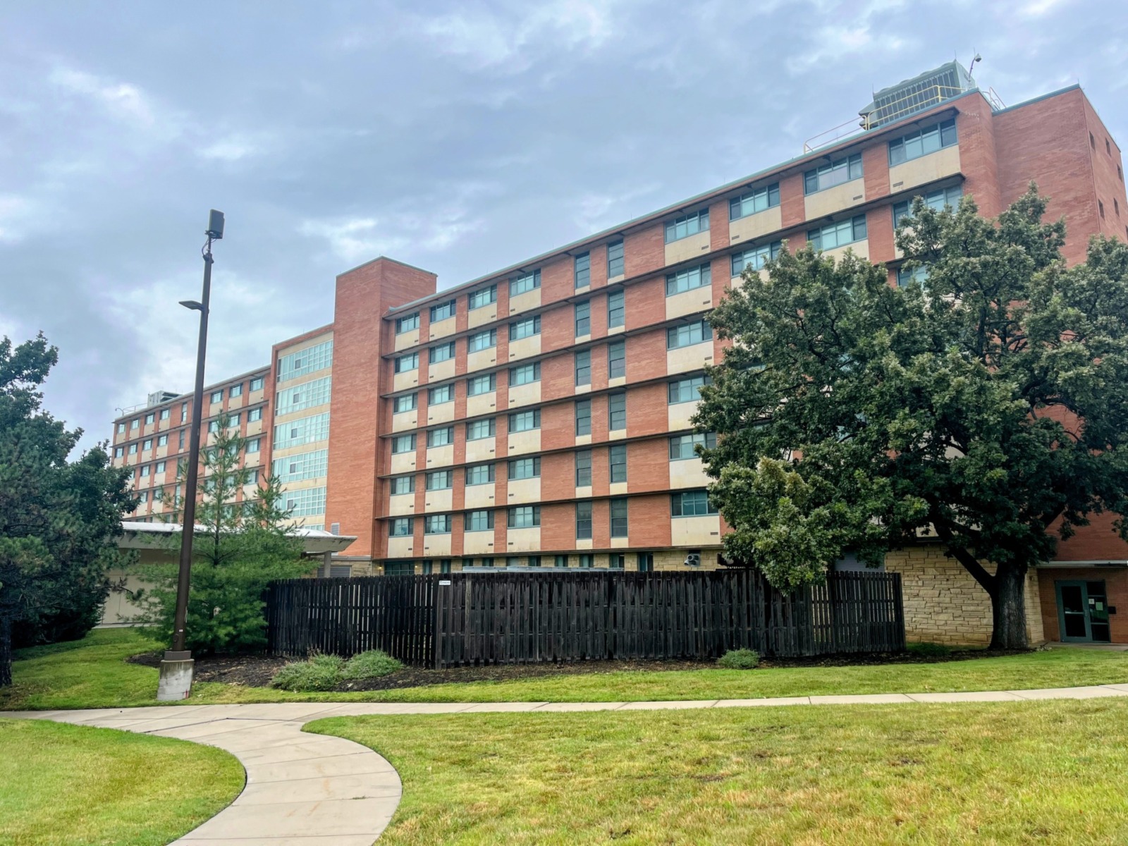 A photo of a dormitory hall. At the foreground of the photo is a green lawn with a curved sidewalk path cutting through it on the left side of the photo. The dormitory is a red color and stretches horizontally across the photo. A large tree partially covers the right side of the building.