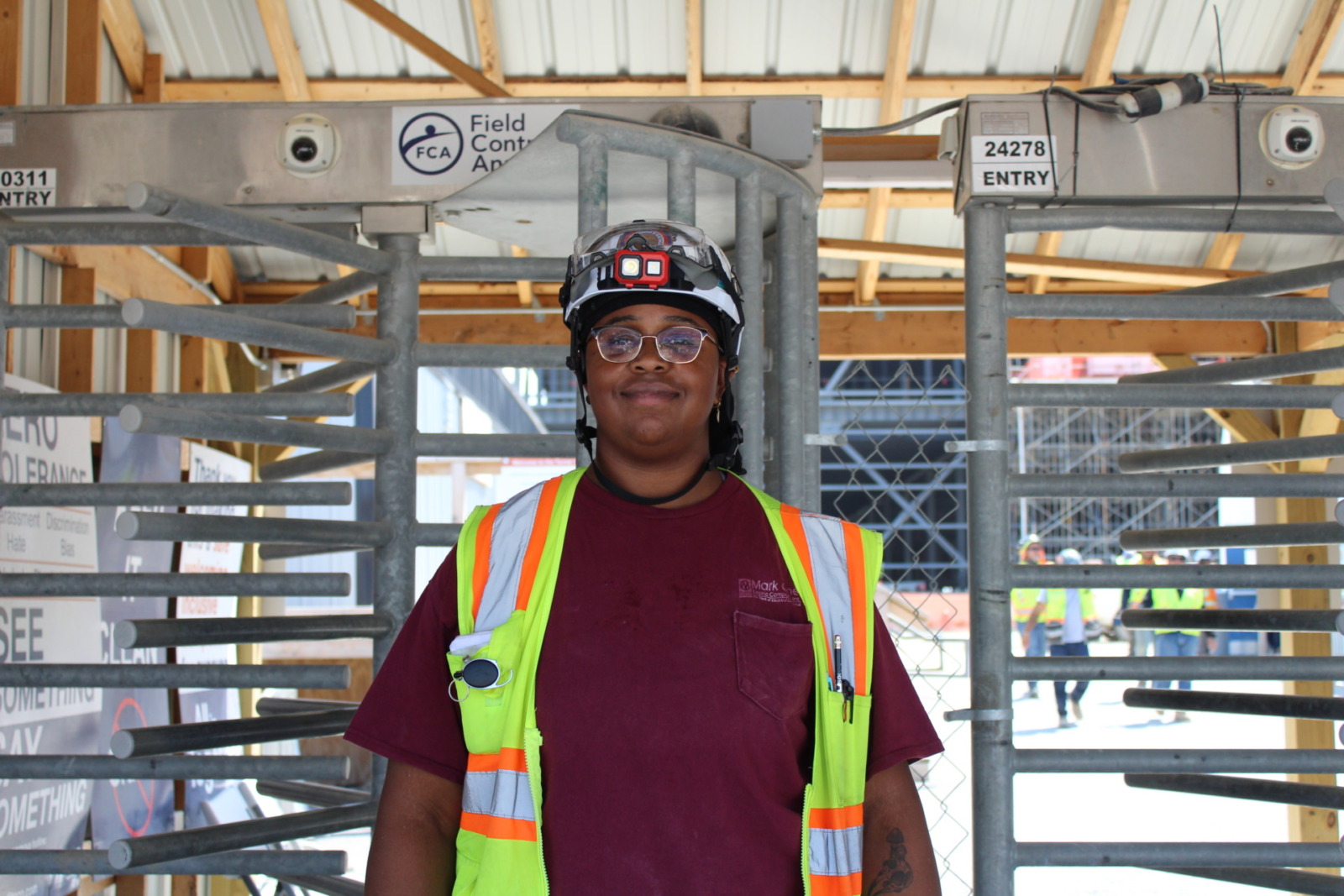 A woman wearing a dark red shirt stands in front of gray metal bars. The woman is wearing glasses, a bright yellow utility vest and a white hard hat.