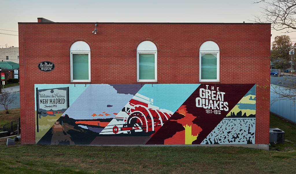 A colorful mural on the side of a brick building with the words "Welcome to Historic New Madrid" and "The Great Quakes" at dusk