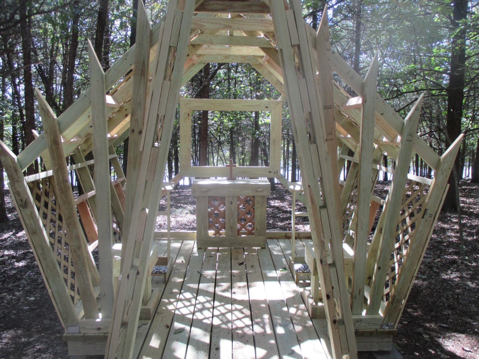 A small wooden Structure shaped like a chapel sits in the woods with dappled sunlight on its floor. Inside is an altar with a cross.