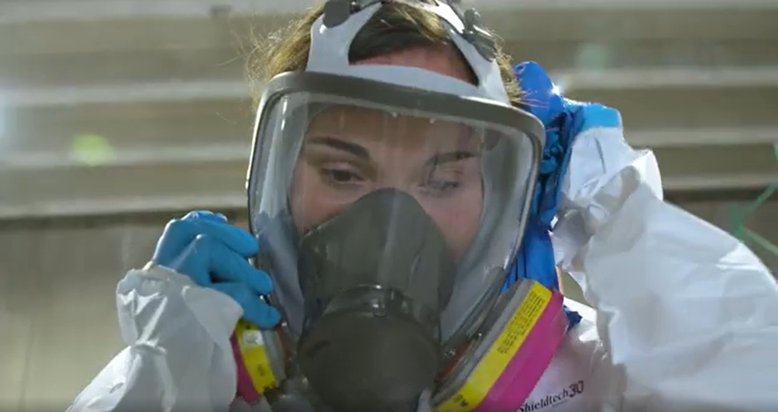 A women in a white hazmat suit puts on a mask.