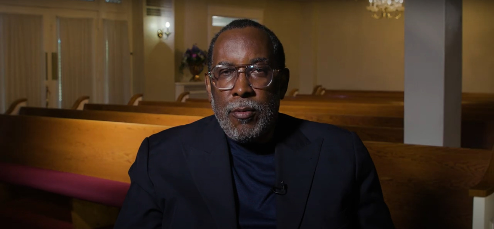A man in a black suit with a beard and glasses sits in a pew in a chapel.