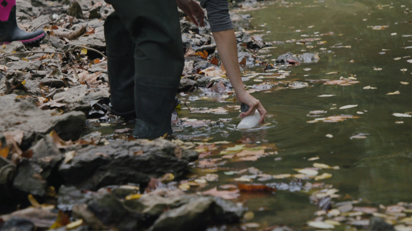 A person bends down and submurges a clear bottle into a green creek. They are wearing black boots. There are orange leaves scattered around them.