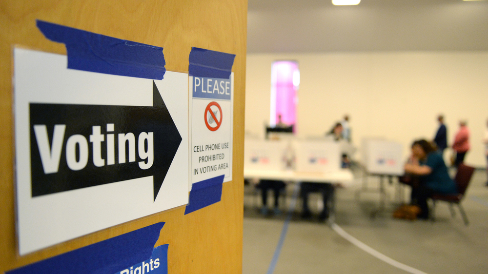 A wooden door opens into a room with blurred figures, white walls and voter dividers sitting on tables. On the door is a sign that has a black arrow and the word Voting. It also has a sign with a red circle with hash through it around a cell phone that says, "cell phone use is prohibited".
