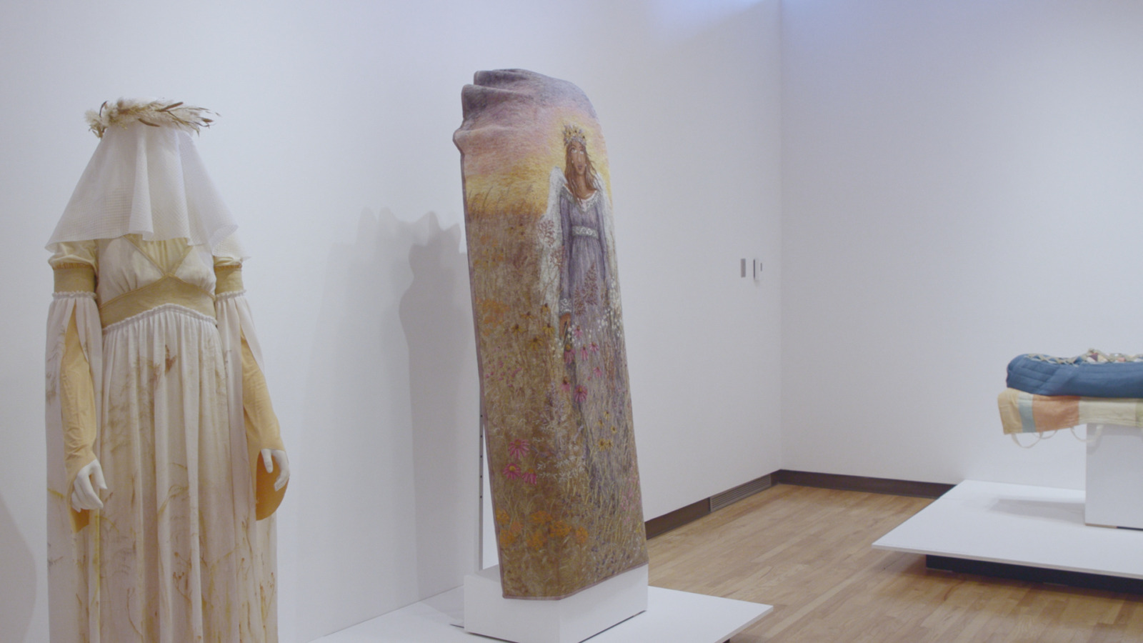 A photo of a museum exhibition. At the left of the photo, a mannequin stands with a light yellow gown and a white veil over its head. The veil is peppered with light brown floral prints. On top of the mannequins head is a crown of prairie grass. To the right of the mannequin with the gown, and in the center of the photo is a coffin cover. The cover depicts an angel in a blue gown with white wings. She is depicted standing in the prairie, covered in prairie flowers. Off to the right side of the photograph is a mannequin lying flat, covered in a blue burial garment.